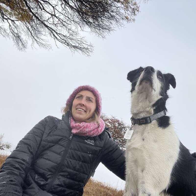 Eine Frau mit rosa Mütze und Schal sitzt draußen neben einem schwarz-weißen Hund. Beide blicken nach oben, umgeben von kahlen Bäumen und einem bewölkten Himmel.