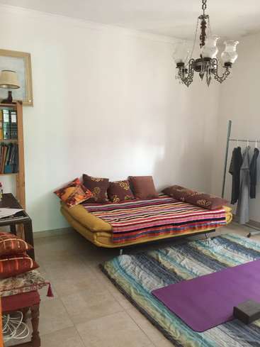 The image depicts a cozy room with a yellow futon, vibrant rug, and eclectic decor, featuring a chandelier, clothing rack, and bookshelf against a crisp white wall.