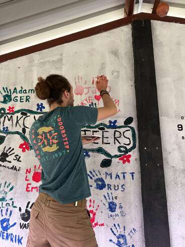 A man with tied-back hair paints his handprint and name on a wall filled with colorful handprints, volunteer names, and cheerful artwork celebrating community involvement.