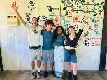 Vier fröhliche junge Leute posieren vor einer Wand, die mit bunten Handabdrücken und Namen bedeckt ist, und feiern ihren ehrenamtlichen Einsatz und ihre Freundschaft, während sie gemeinsam fröhlich lächeln.