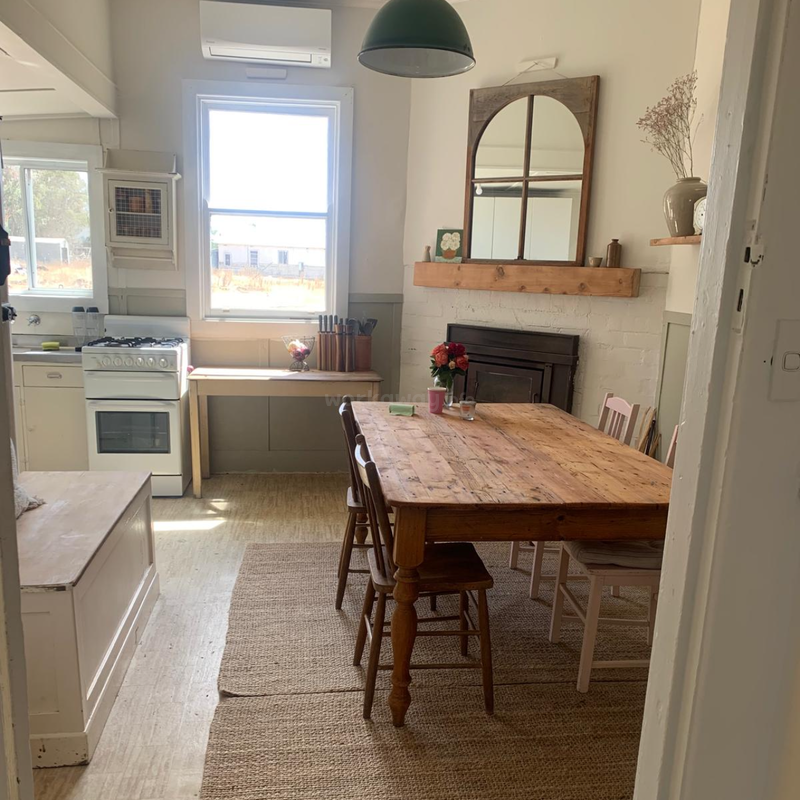 Une cuisine-salle à manger rustique et confortable avec des meubles en bois, une grande table, un poêle, des fleurs, un miroir, une cheminée, de la lumière naturelle et des tons neutres, créant une atmosphère chaleureuse.