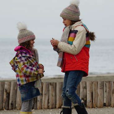 Dos chicas jóvenes están en una playa, con gorros y abrigos de invierno, con una valla de madera y el océano de fondo, transmitiendo una sensación de amistad y alegría.