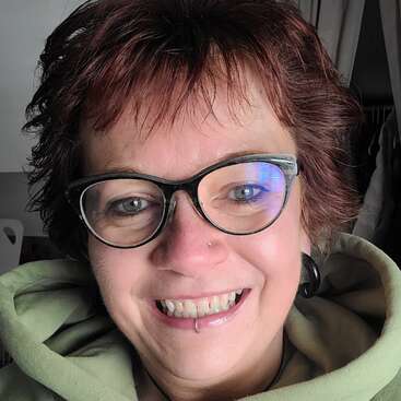 Una persona alegre, con el pelo corto castaño rojizo, gafas y una sudadera con capucha verde, sonríe con entusiasmo. Lleva un piercing en la nariz y otro en el labio.