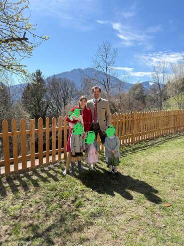 Uma família de cinco pessoas está ao ar livre, em um gramado verde ao lado de uma cerca de madeira. No fundo, montanhas e árvores sob um céu azul brilhante.