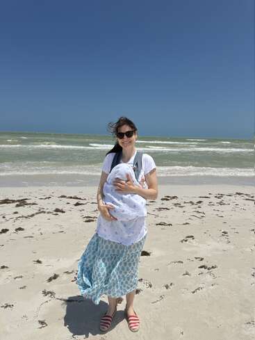 Uma mulher sorridente, usando óculos escuros, está em uma praia ensolarada segurando um bebê enrolado em um cobertor. As ondas do mar e o céu azul claro criam um cenário sereno.
