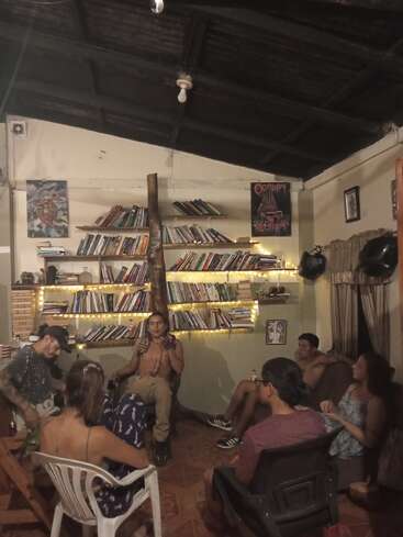 Sechs Personen sitzen in einem gemütlichen, schwach beleuchteten Raum voller Bücher und Lichterketten und unterhalten sich. Die Atmosphäre ist entspannt, informell und einladend.