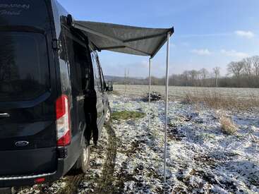 Ein schwarzer Wohnwagen mit einer großen Markise steht auf einem verschneiten Feld, umgeben von Bäumen, unter einem blauen Himmel mit ein paar Wolken. Eine ruhige Wildnis.