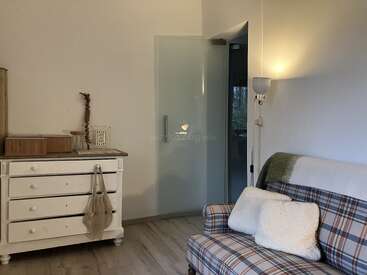 Ein gemütliches Zimmer mit einem karierten Sofa, weißen Kissen, einer Decke, einer weißen Kommode mit Deko, Holzboden, Glastür und sanfter Wandbeleuchtung.