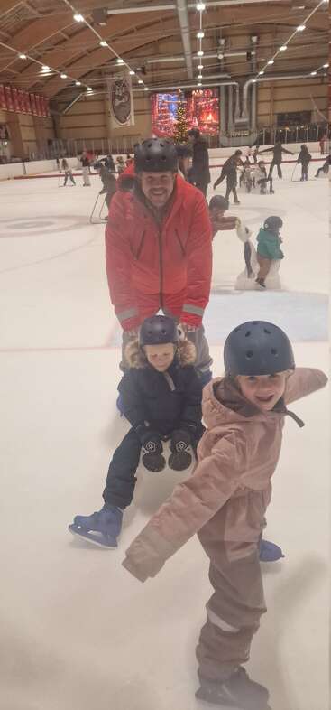 Un hombre y dos niños, todos con cascos y ropa de invierno, disfrutan patinando sobre hielo en una pista cubierta. Al fondo se ven otros patinadores y decoraciones navideñas.