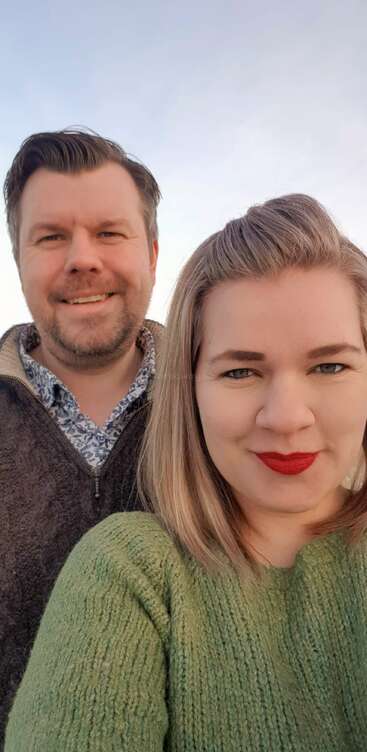 La imagen muestra una fotografía de dos individuos, probablemente una pareja, de pie juntos, con la persona de la izquierda vistiendo una camisa oscura y la persona de la derecha vistiendo una camisa de color claro.