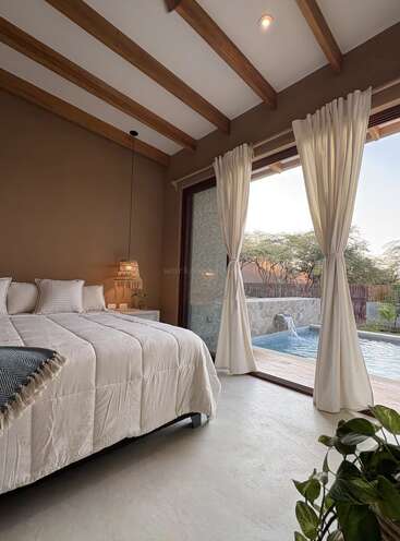 Um quarto aconchegante com tetos com vigas de madeira, roupa de cama branca, iluminação suave e grandes portas de vidro com cortinas esvoaçantes que se abrem para uma piscina tranquila e uma vista exuberante do exterior.
