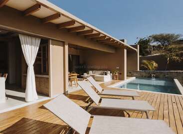 Um pátio moderno e bem iluminado, com deck de madeira, conta com três espreguiçadeiras, uma pequena piscina, áreas confortáveis para refeições e descanso ao ar livre, cortinas brancas e árvores frondosas ao redor.