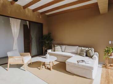 Este pátio aconchegante tem um sofá modular branco, uma poltrona, uma mesinha de centro, vasos de plantas e paredes em tons quentes de bege com vigas de madeira à vista, tudo banhado pela luz natural do sol.