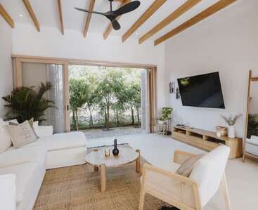 Essa sala de estar bem iluminada tem um sofá modular branco, vigas de madeira, plantas de interior, uma mesa de centro redonda, TV, decoração moderna e grandes portas de vidro que se abrem para a vegetação.