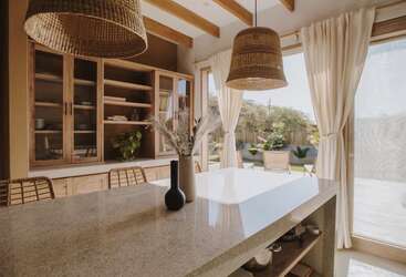A cozinha bem iluminada tem uma ilha de mármore, luminárias suspensas de rattan, cadeiras de vime, armários de madeira, cortinas brancas e vista para o jardim, criando um ambiente aconchegante e convidativo.