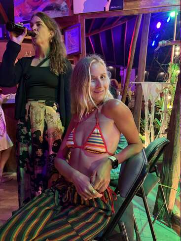 Duas mulheres estão em um espaço interno animado e colorido. Uma delas está sentada, sorrindo, vestindo um biquíni listrado e calças, enquanto a outra está em pé, bebendo uma cerveja. Há obras de arte decorando o fundo.