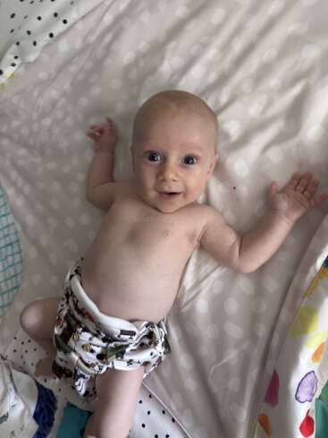 Um bebê sorridente está deitado sobre um cobertor macio e estampado, usando uma fralda de pano. O bebê parece feliz e brincalhão, com os braços estendidos e os olhos brilhantes.