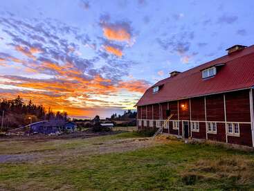 Das Bild zeigt eine ruhige ländliche Szene bei Sonnenuntergang mit einer großen roten Scheune mit rotem Dach und weißer Zierleiste vor einem leuchtend orangefarbenen und lila Himmel.