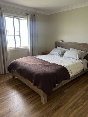 A cozy bedroom features a wooden bed with white bedding and a brown blanket, wooden floor, light curtains, a window, and a small bedside table.