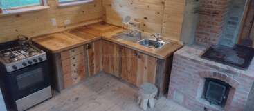Cette cuisine chaleureuse est dotée d'armoires en bois, d'un évier en métal, d'un petit four avec une cuisinière, d'un four en brique, d'un tabouret rustique, ainsi que de murs et de sols en bois, créant ainsi une atmosphère chaleureuse.
