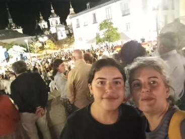 Duas mulheres sorriem em primeiro plano, tirando uma selfie em um animado evento noturno ao ar livre. Muitas pessoas e edifícios históricos são visíveis ao fundo.