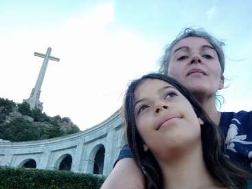 Uma mulher e uma menina tiram uma selfie juntas em frente a uma grande cruz de pedra e a uma estrutura em arco, semelhante a um monumento, sob um céu azul.