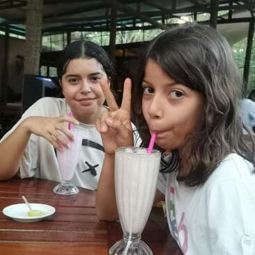 The image shows two young girls sitting at a table with milkshakes, one making a peace sign with her hand. They are wearing white shirts.