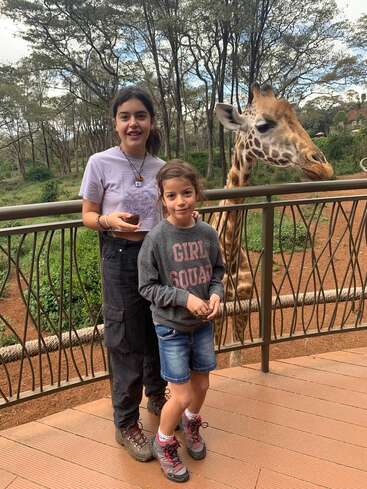 A imagem mostra duas meninas em pé atrás de uma grade, com uma girafa visível ao fundo, em um cenário de árvores e arbustos.