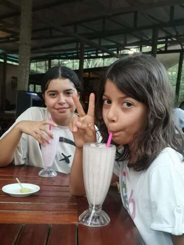 Das Bild zeigt zwei junge Mädchen, die an einem Tisch mit Milchshakes sitzen, wobei die eine mit ihrer Hand ein Friedenszeichen macht. Sie tragen weiße Hemden.