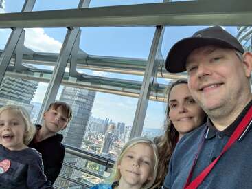 Uma família de cinco pessoas sorri para uma selfie dentro de um prédio de vidro com arranha-céus da cidade e céu azul claro visível pelas janelas ao fundo.