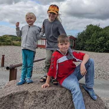 Tres niños sonrientes posan juntos al aire libre sobre una gran roca. El fondo muestra un suelo cubierto de guijarros, vegetación, un edificio rojo y un cielo nublado.