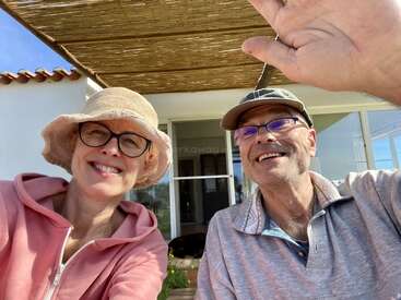 A smiling man and woman wearing glasses, hats, and casual clothes sit outside under a shaded patio, enjoying a sunny day. The man waves at the camera.