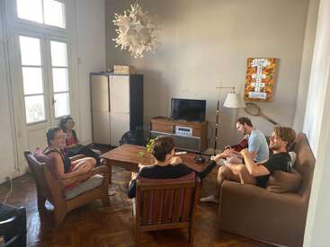 Five friends are relaxing and socializing in a cozy living room. One is playing guitar, while others sit chatting and enjoying time together. Sunlight brightens space.