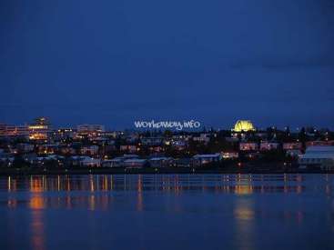 The image depicts a cityscape at night, with a body of water in the foreground and a large building with a lit-up dome in the distance.