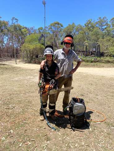 Deux personnes portant des équipements de protection et des casques se tiennent à l'extérieur sur de la terre, entourées d'arbres. L'une d'elles tient une tronçonneuse, tandis qu'un sac à dos et une corde sont posés à proximité. Le temps est ensoleillé.