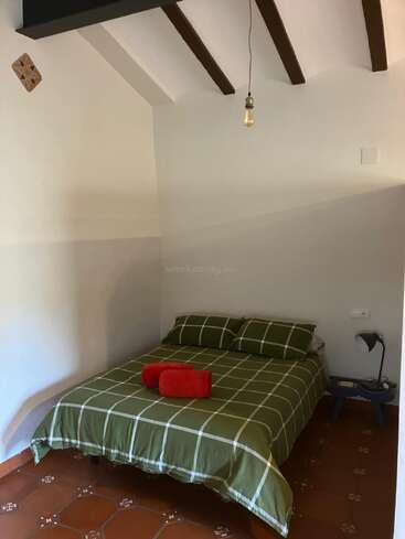 This image shows a simple bedroom with a green checkered bedspread, two red towels, a small bedside table, a lamp, and an exposed lightbulb.