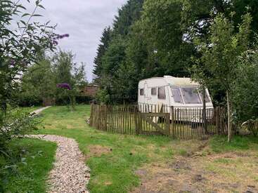A white caravan is parked inside a wooden fenced area surrounded by green trees, grass, and a gravel path, creating a peaceful countryside setting under cloudy skies.