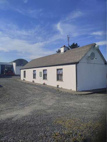 A small white house with a grey roof sits on a gravel lot, with potted plants, bright blue sky, and farm structures and tractor in the background.