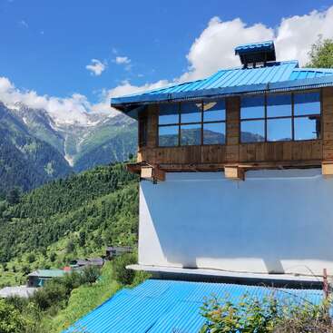 Una casa con tejado azul y grandes ventanales domina exuberantes colinas verdes y lejanas montañas nevadas bajo un cielo azul brillante lleno de nubes esponjosas.