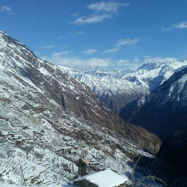 La imagen representa un sereno paisaje montañoso con picos nevados y campos en terrazas, sobre un cielo azul claro con nubes dispersas.