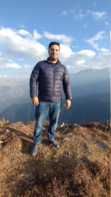 A man stands confidently on a grassy hilltop, wearing a blue jacket and jeans, with mountains and a partly cloudy sky in the background.