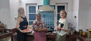 Three women are smiling in a kitchen. One is holding a tray of vegetables, another stands in the center, and the third is holding a spatula, ready to cook.