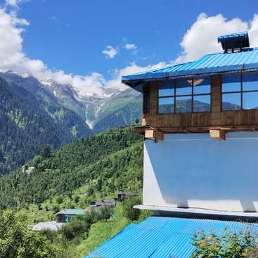 A house with blue roof and large windows overlooks lush green hills and distant snow-capped mountains under a bright blue sky filled with fluffy clouds.