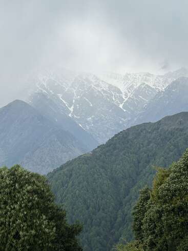 Derrière des collines verdoyantes, se profilent de majestueuses montagnes aux sommets enneigés, enveloppées de nuages brumeux. Des forêts denses recouvrent les pentes inférieures, créant un paysage naturel empreint de sérénité.