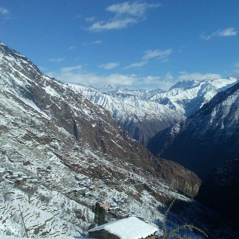 The image depicts a serene mountainous landscape with snow-covered peaks and terraced fields, set against a clear blue sky with scattered clouds.