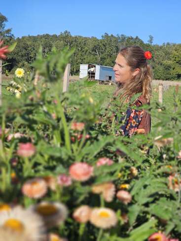 Una mujer con una flor en el pelo está de pie en un soleado jardín rodeado de coloridas flores en flor, con árboles y un pequeño cobertizo al fondo.