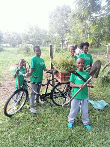 Cinq enfants vêtus de chemises vertes sourient à l'extérieur, debout à côté d'un vélo noir équipé d'un panier rempli de plantes. Ils tiennent des outils de jardinage, entourés d'herbe verte et d'arbres.