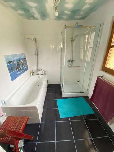 This bathroom has a bathtub, separate glass shower, dark tiled floor, blue mat, wooden stool, towel rack, ocean mural, and sky-themed painted ceiling.