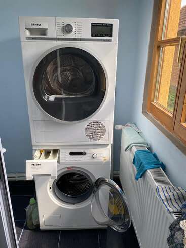A washing machine and dryer are stacked in a small laundry room with a window. Detergent bottles and cloths are visible on a radiator nearby.