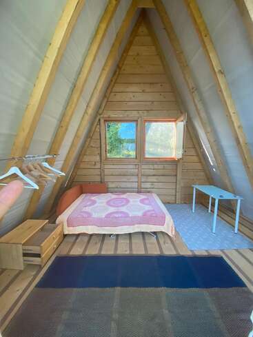 This cozy, triangular wooden cabin features a simple bed with a colorful quilt, a small table, window, clothes rack, wooden drawers, and two rugs.